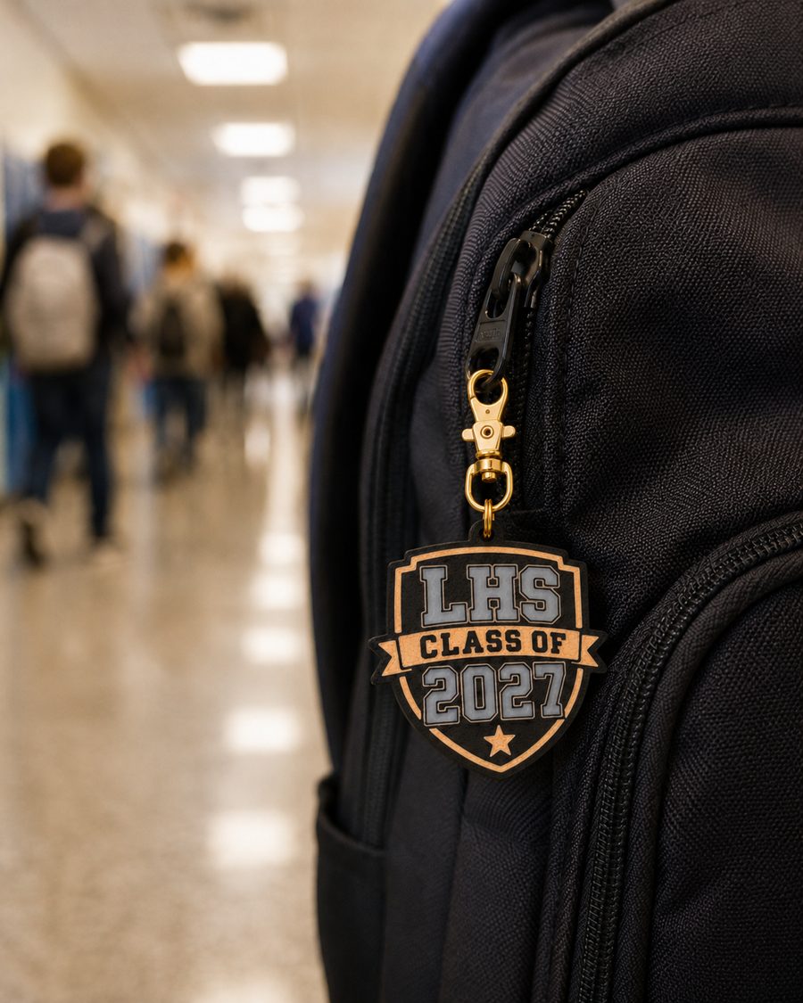 LHS Class of 2027 shield bag charm clipped to a black backpack in a school hallway