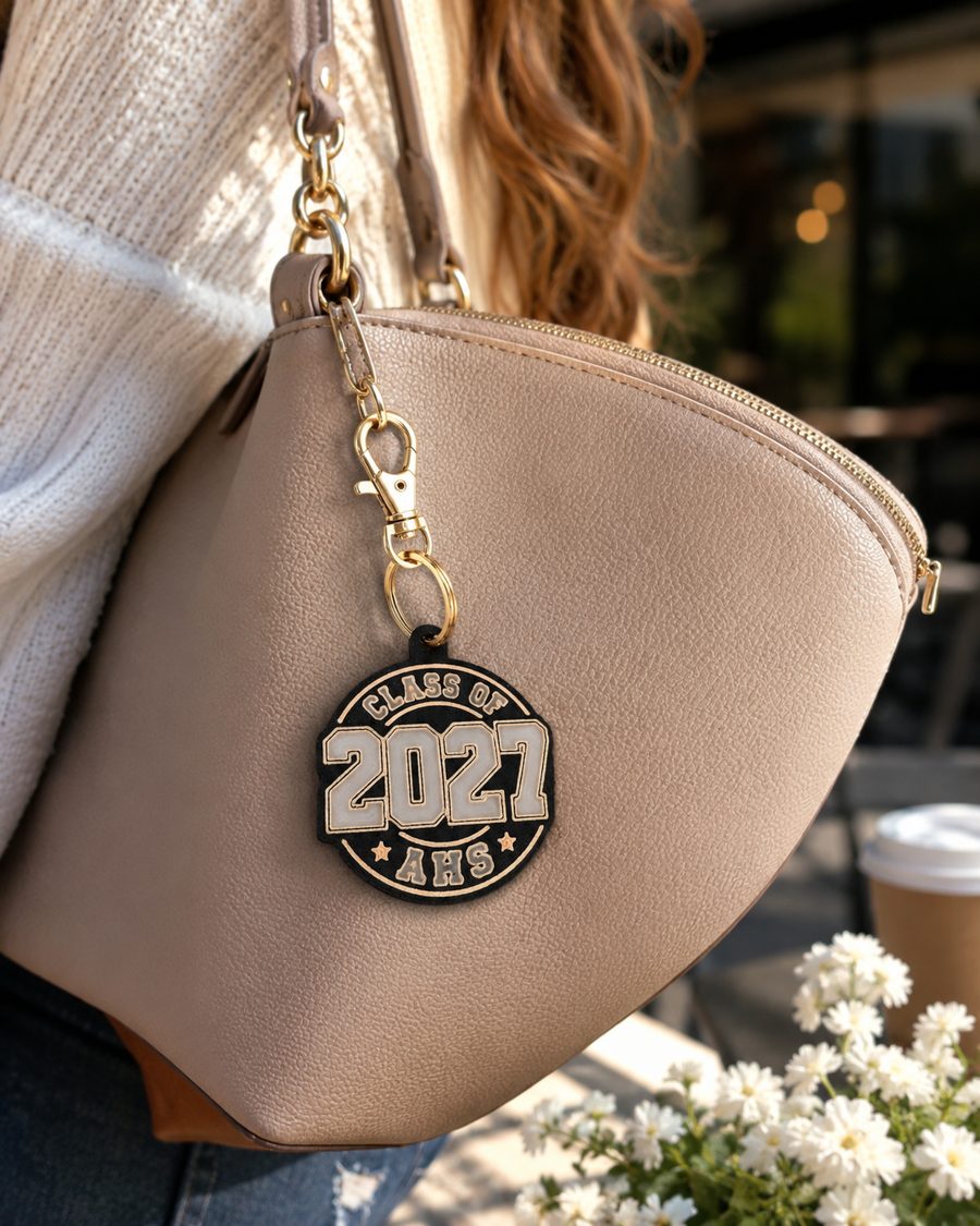 AHS Class of 2027 round bag charm in black and gold clipped to a tan purse